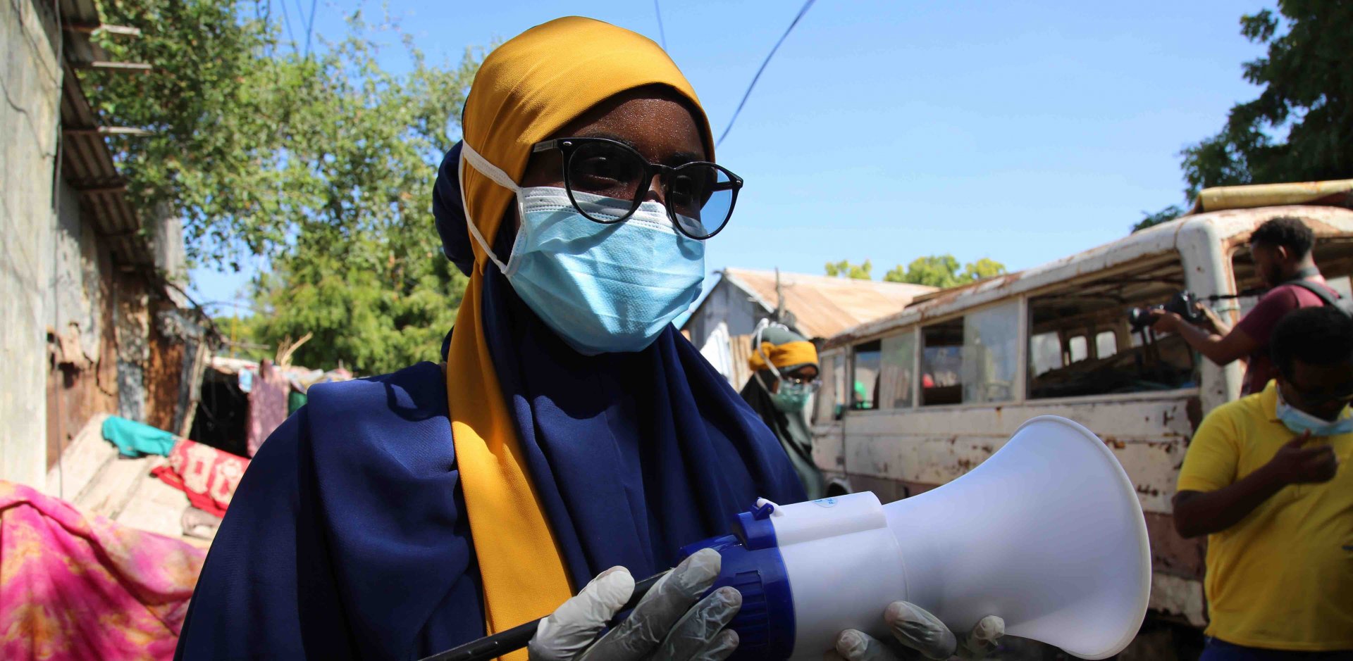 A Somali woman wearing a yellow headscarf, black framed glasses and a medical face mask holds a bullhorn